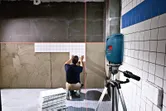Person aligns wall tiles using a laser leveling tool set on a tripod.
