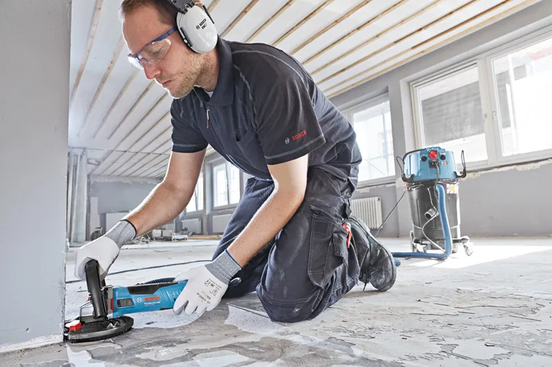 A person wearing safety equipment grinds a concrete floor with a blue concrete grinder.