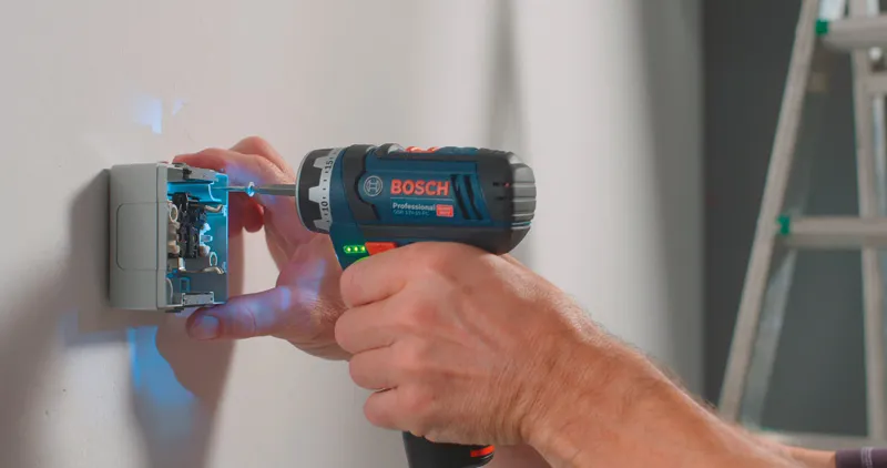 A person uses a cordless drill driver to fasten screws into an electrical box on a wall.