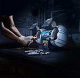 A person wearing safety equipment sharpens a drill bit with a double-wheeled bench grinder.