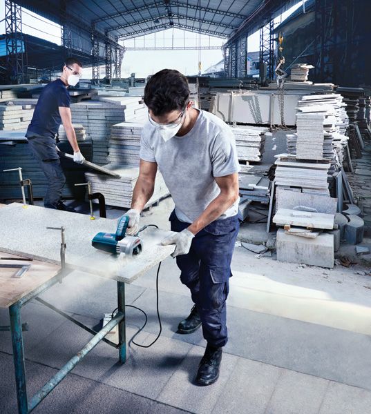 A person wearing safety equipment cuts a stone slab with a power saw in a warehouse.