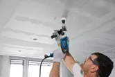 A person wearing safety equipment drills a circular hole into a ceiling with a power tool.