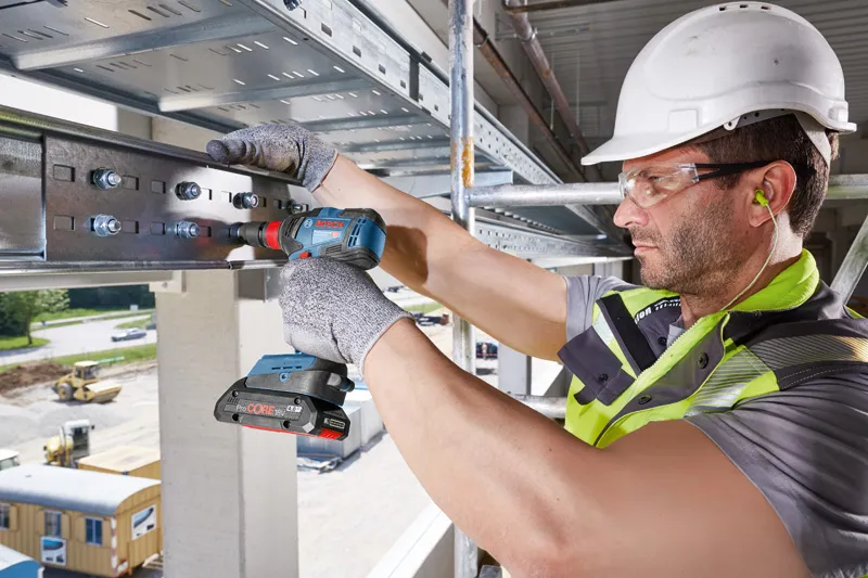 A person wearing safety equipment uses a cordless drill to secure bolts on a steel beam.
