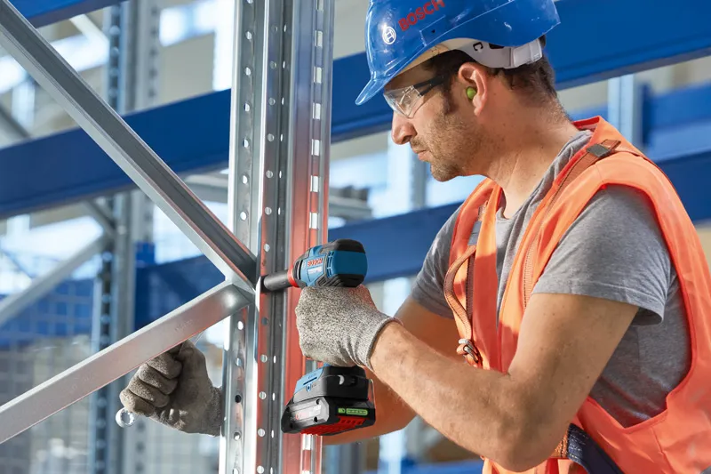 A person wearing safety equipment secures a metal frame with a cordless power drill.