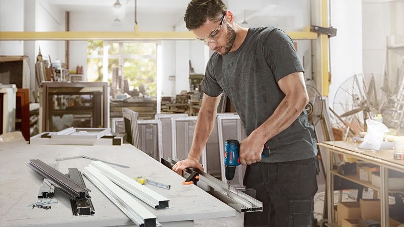 A person wearing safety equipment drills into a metal profile at a workbench.