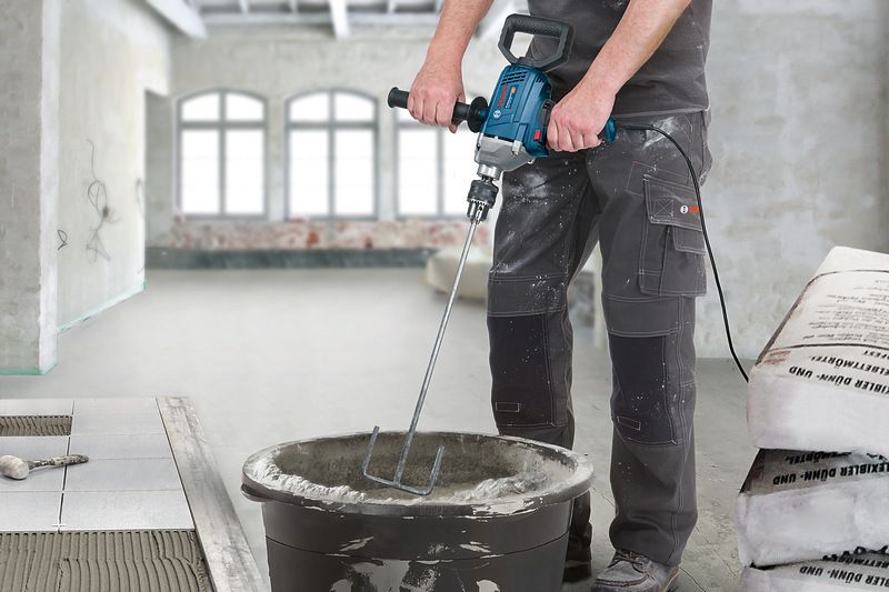 A person wearing safety equipment mixes cement in a bucket using a power mixer.