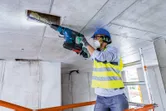 A person wearing safety equipment drills into a concrete ceiling with a cordless rotary hammer.
