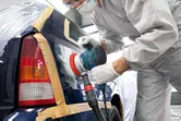 A person wearing safety equipment sands a car’s rear panel with a random orbit sander.