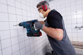 A person wearing safety equipment drills into bathroom wall tiles with a cordless rotary hammer.