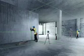 Two workers wearing safety equipment align interior walls using a laser leveling tool.