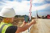 A person wearing safety equipment uses a laser receiver on a leveling rod at a construction site.