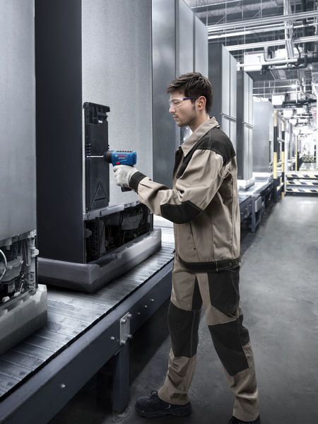 A person wearing safety equipment uses a power tool to inspect equipment on an assembly line.
