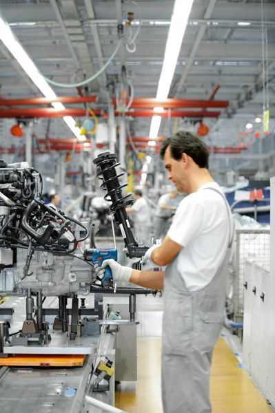 Person wearing safety equipment uses a power tool to assemble machinery on a factory line.