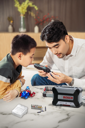 An adult and a child examine a cordless screwdriver with tool bits on a table.