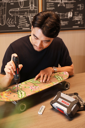 Person using a cordless screwdriver to repair a floral skateboard on a table.