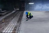 A person wearing safety equipment scans concrete with a detector near exposed rebar.