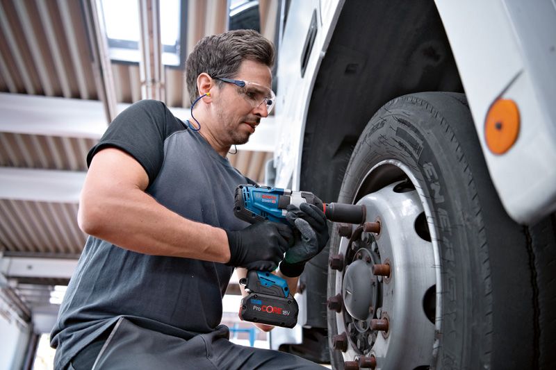 A person wearing safety equipment uses a cordless impact wrench on a truck wheel.