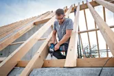 A person wearing safety equipment fastens a wooden beam with a cordless impact wrench.