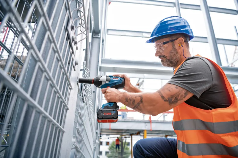 A worker wearing safety equipment uses a cordless impact wrench on a metal fence.
