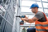 A worker wearing safety equipment uses a cordless impact wrench on a metal fence.