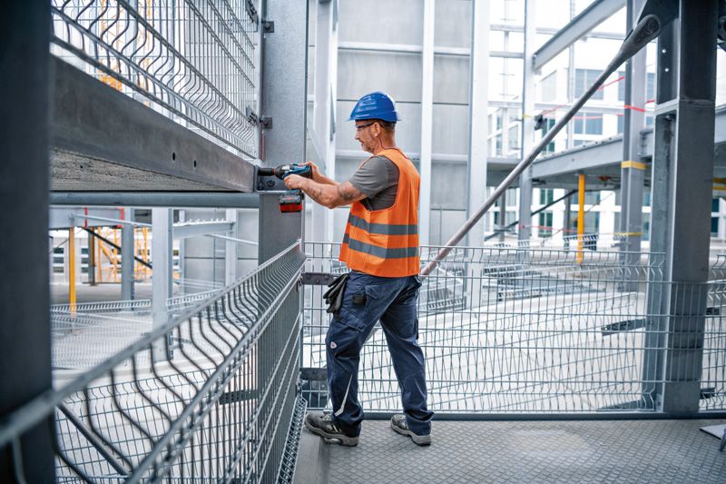 A person wearing safety equipment fastens a bolt to a steel beam with an impact wrench.