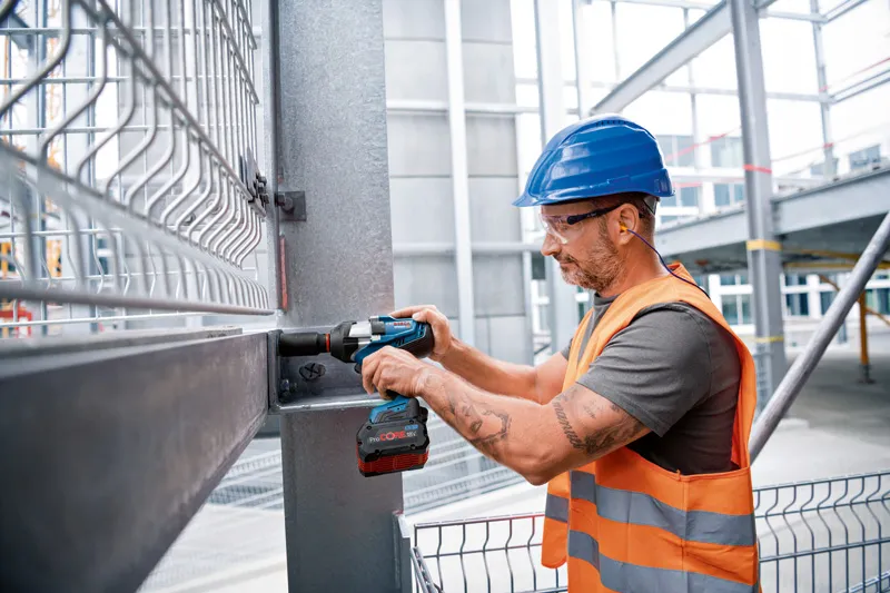 A person wearing safety equipment uses a cordless impact wrench on a steel beam.