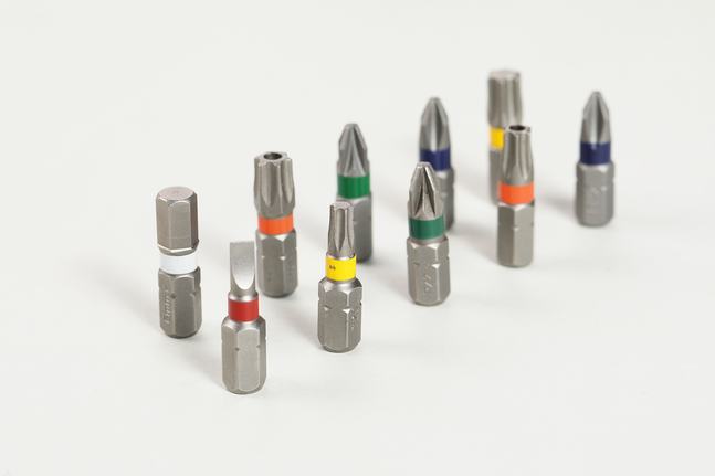 Assorted screwdriver bits with colored bands are arranged on a light surface.