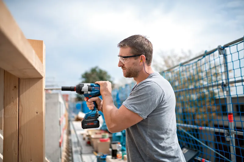A person wearing safety equipment uses a cordless impact wrench on a wooden beam.