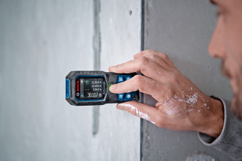 A person measures a wall with a laser measure, displaying several distance readings.