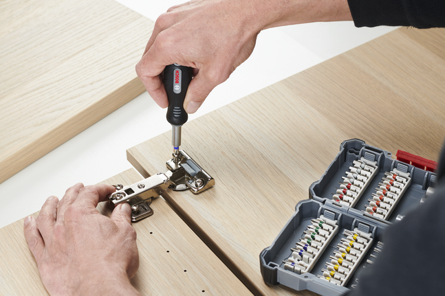 Person assembling a cabinet hinge on light wood near a case of assorted screwdriver bits.