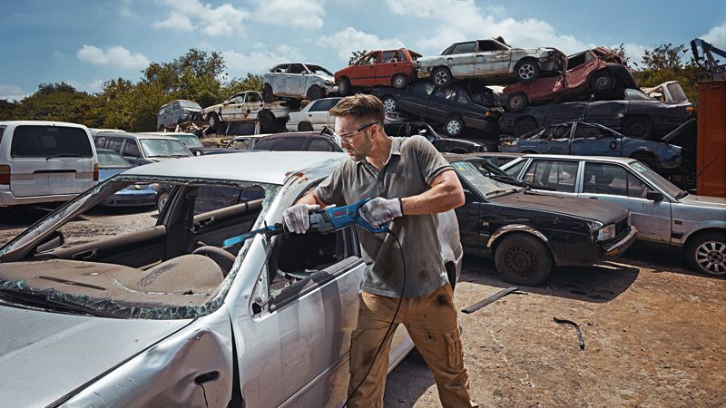 A person wearing safety equipment cuts a car door with a reciprocating saw in a scrapyard.