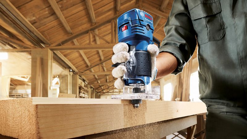 A person wearing safety equipment uses a palm router to shape the edge of a wooden board.