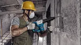 A person wearing safety equipment uses a demolition hammer to break a concrete wall.