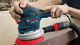 A person uses a random orbit sander to smooth the edge of a wooden surface.
