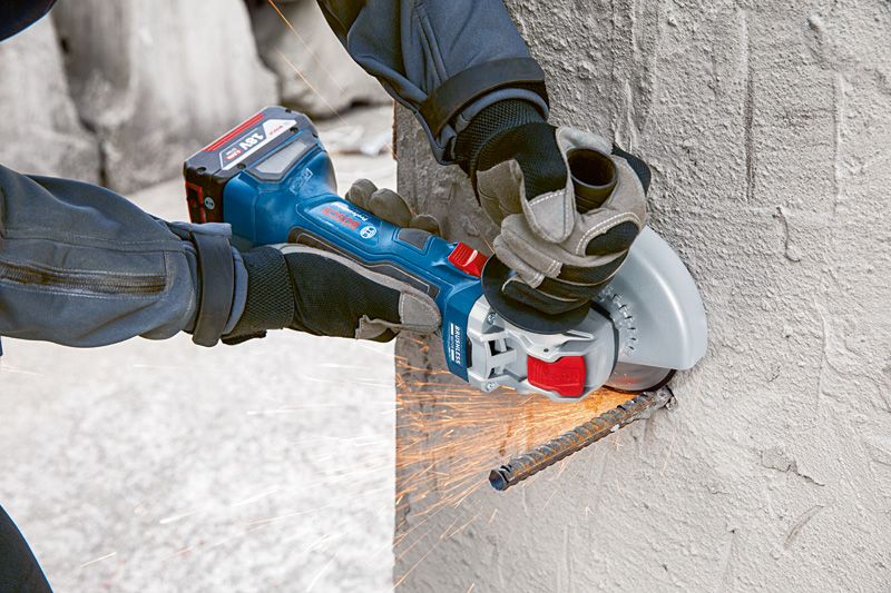 A person wearing safety equipment uses a cordless angle grinder to cut rebar in concrete.