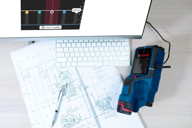 Digital wallscanner detector on architectural blueprints beside a computer keyboard.