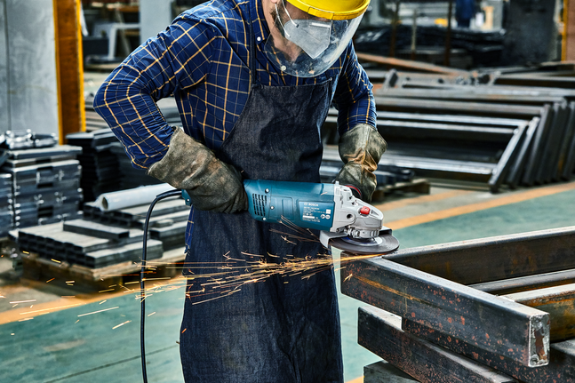 Person wearing safety equipment grinds metal beams, creating sparks in a workshop.