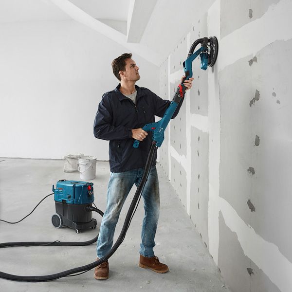 A person sands drywall with a drywall sander connected to a dust extractor.