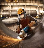 A person wearing safety equipment grinds a large metal pipe with sparks flying.