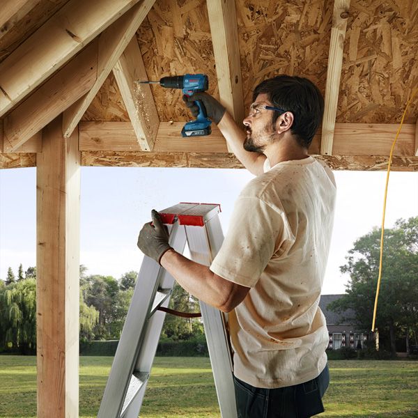 A person wearing safety equipment drills into a wooden beam while standing on a ladder.