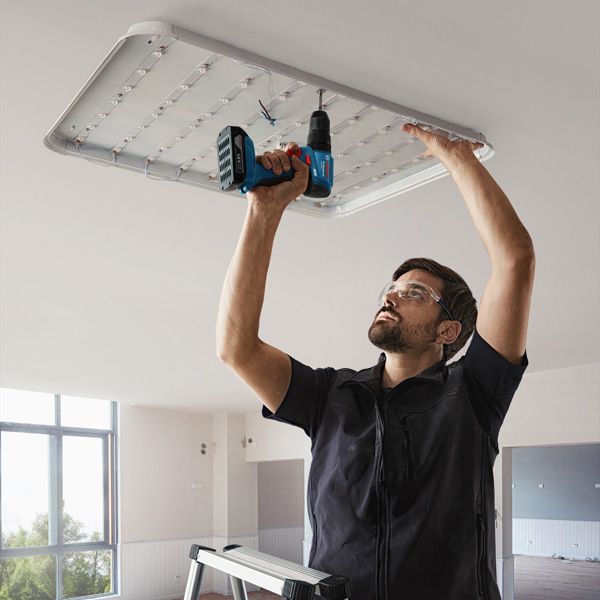 A person wearing safety equipment installs a large ceiling light using a cordless drill.