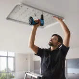 A person wearing safety equipment installs a large ceiling light using a cordless drill.