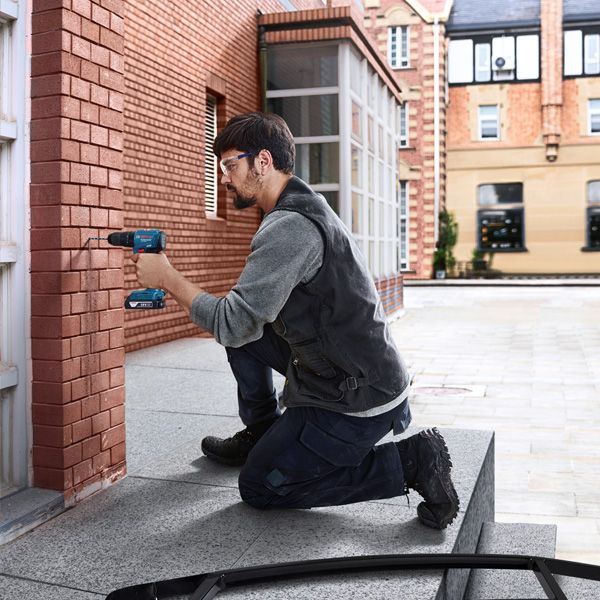 Person wearing safety equipment drills into a brick wall outdoors.