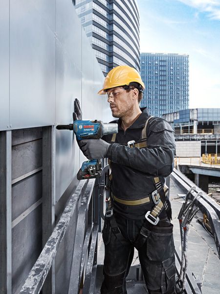 Person wearing safety equipment drills into a metal panel on a building exterior.