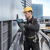 A person wearing safety equipment uses a power drill to fasten panels on a building exterior.