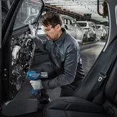 A person wearing safety equipment uses a power tool to assemble a car door in a factory.
