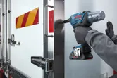 A person wearing safety equipment uses a cordless impact driver on a metal truck door.