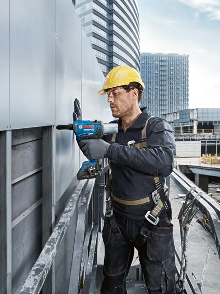 A person wearing safety equipment fastens metal siding using a cordless rivet gun.