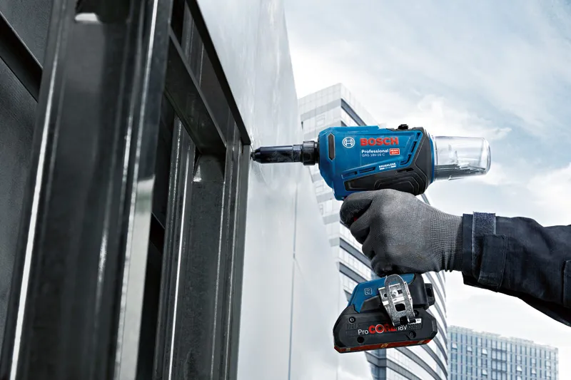 A person wearing safety equipment fastens a metal panel with a cordless rivet gun.