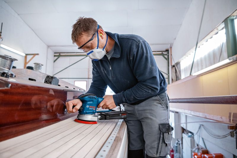 A person wearing safety equipment sands a wooden boat deck using a random orbit sander.